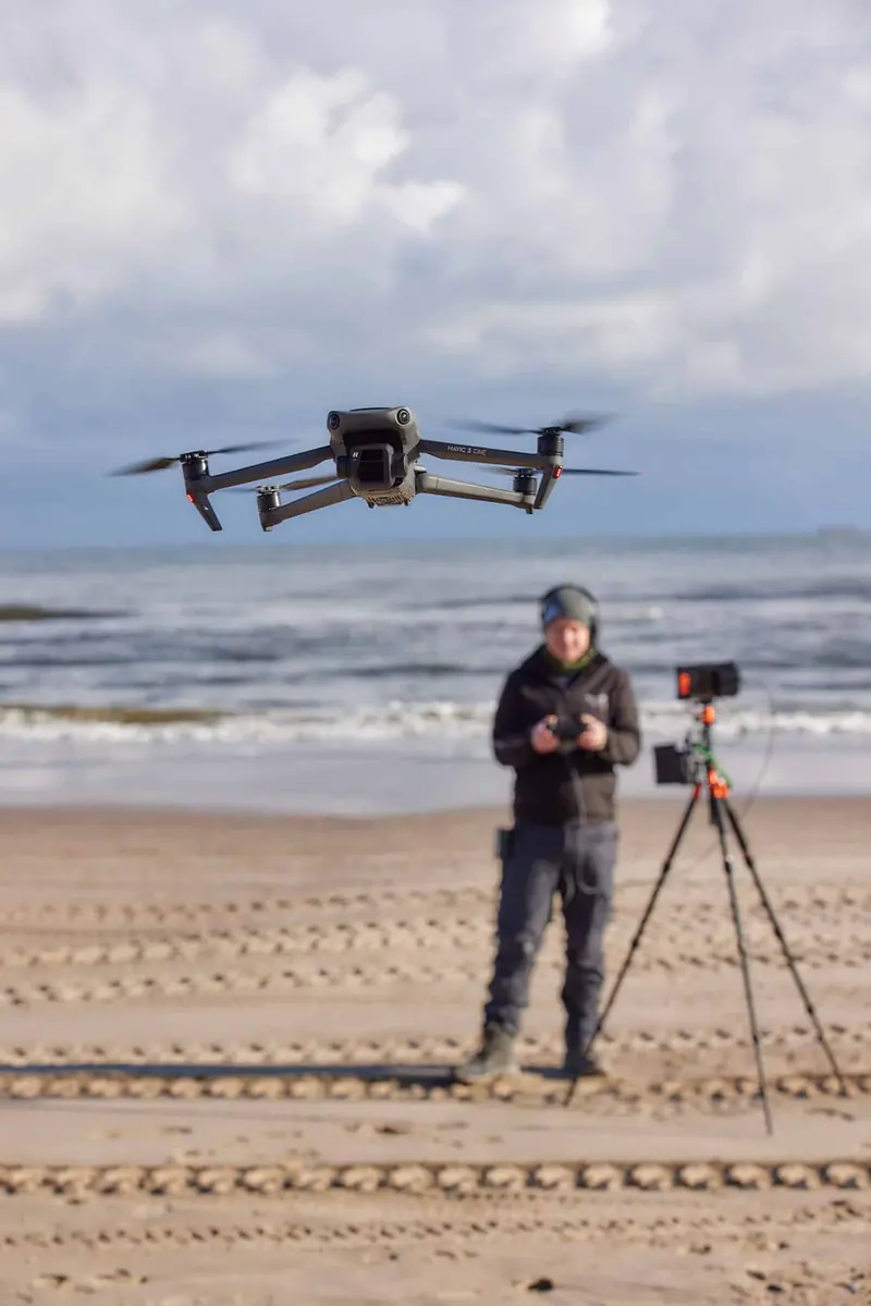 Kameradrohne im Vordergrund, dahinter der Pilot mit Fernsteuerung am Strand